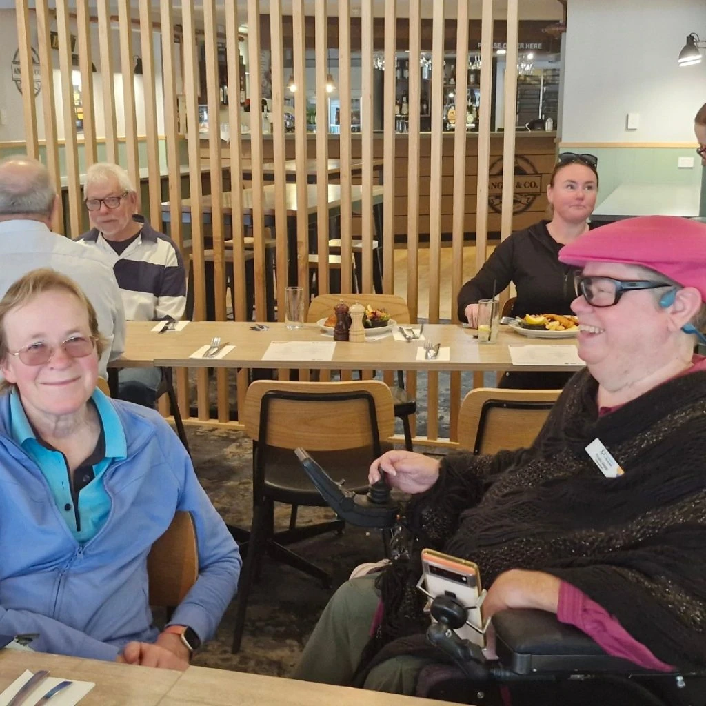 Two people sit at a table in a restaurant, one in a wheelchair. Both are smiling. Other patrons can be seen in the background, seated at different tables.