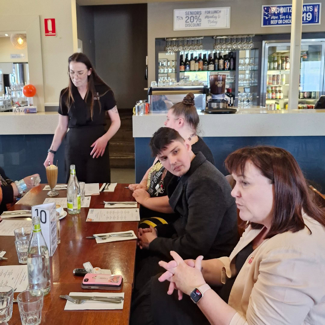 A group of people seated at a restaurant table engage in conversation. A waitress, standing, takes their orders. Menus, drinks, and a table number 10 are visible on the table.