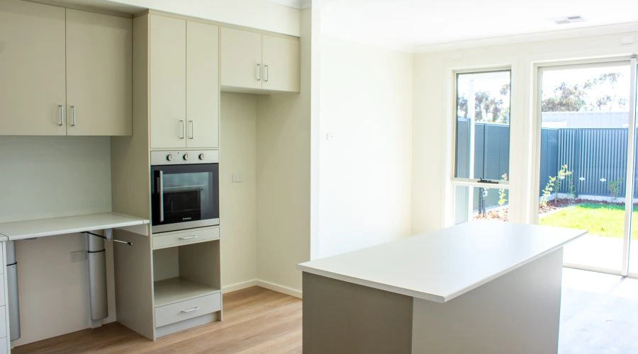 Modern kitchen with light-colored cabinets, an oven, and a central island. Large windows provide natural light, and a glimpse of a grassy backyard is visible.