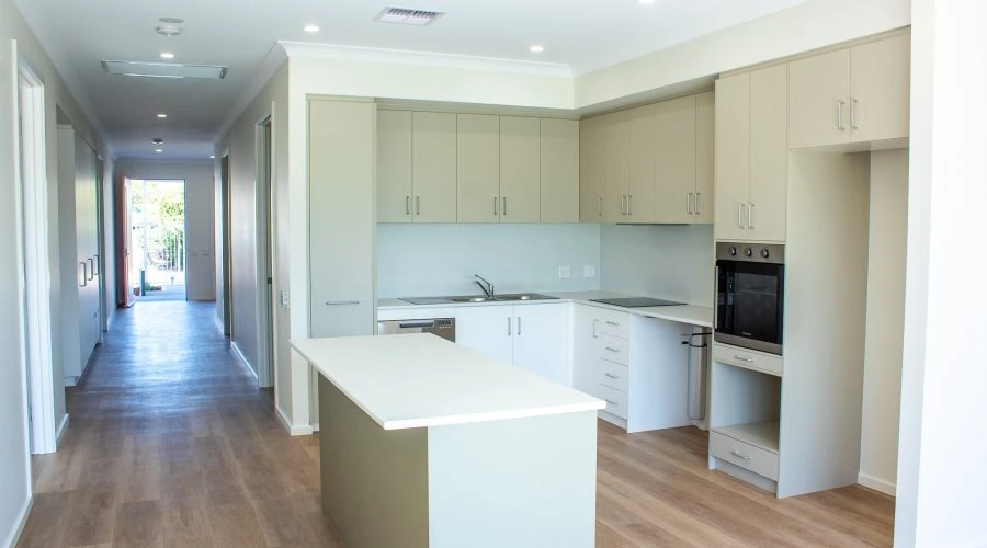 Modern kitchen interior with beige cabinets, a white island, built-in oven, and light wood flooring. A hallway leads to a doorway with natural light.