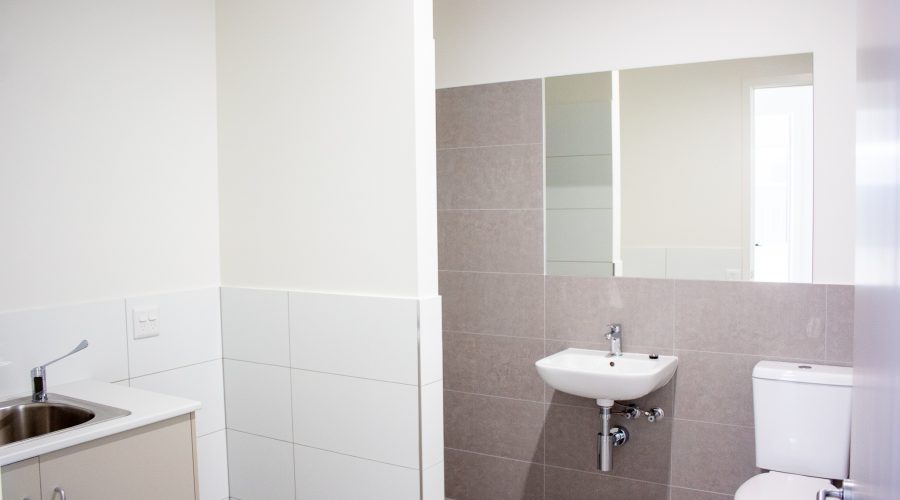 Minimalist bathroom with white and beige tiles, featuring a sink, toilet, and wall mirror. A small countertop with a sink is on the left.