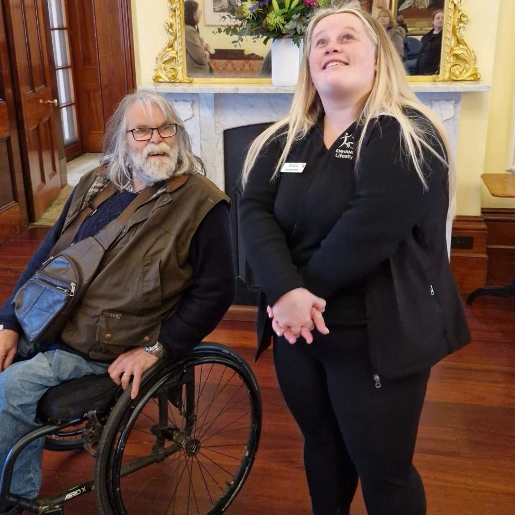 A man in a wheelchair with long hair and glasses sits next to a standing woman in a black uniform. They are indoors in front of a fireplace.