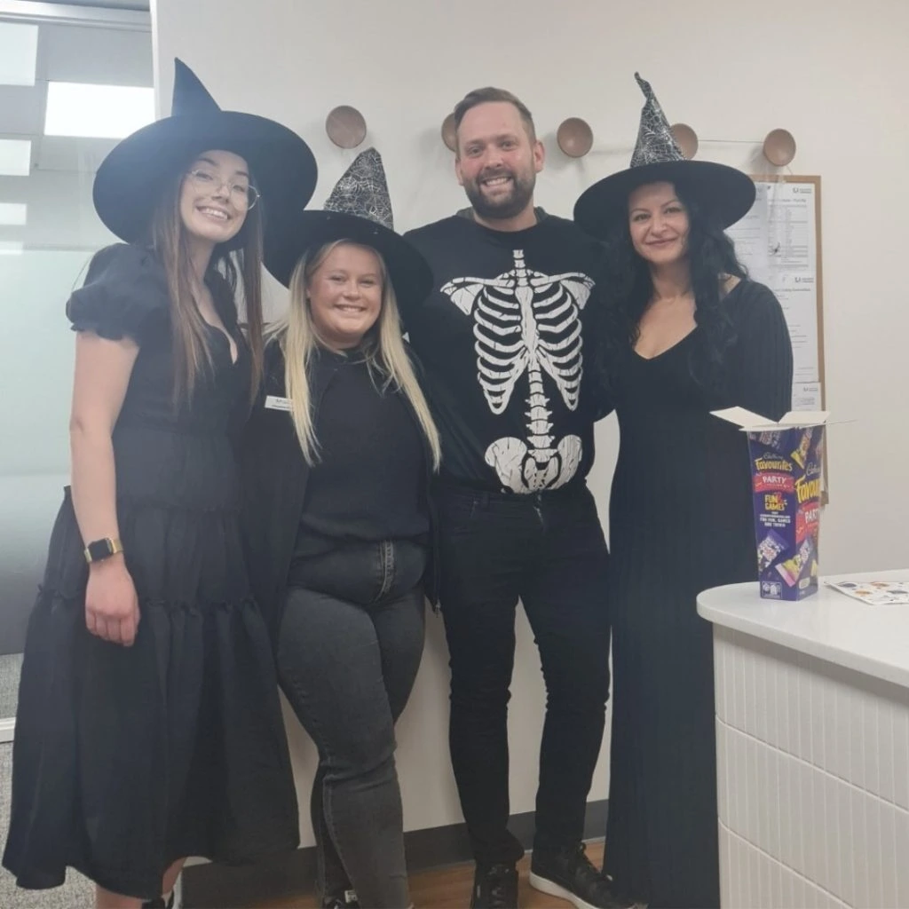 Four people in Halloween costumes, three dressed as witches and one wearing a skeleton shirt, stand smiling in an office setting.