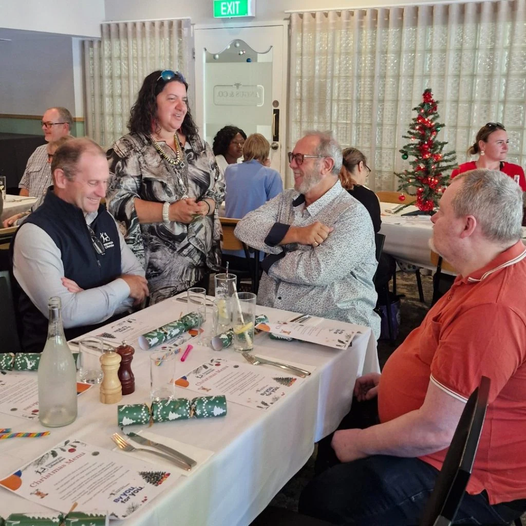 A group of people sits and stands around a dining table during a holiday gathering. Christmas tree decorations are visible in the background.