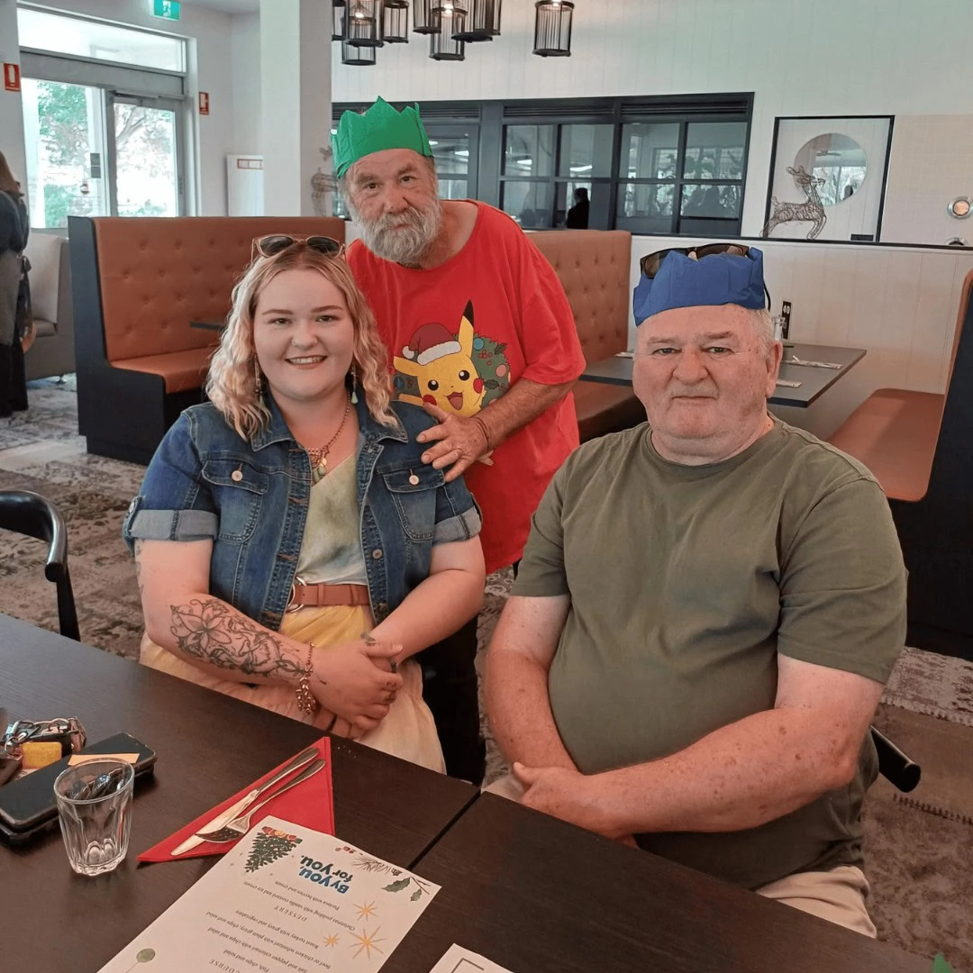 Three people wearing festive paper crowns are sitting and standing around a table in a restaurant. One person is standing behind the other two, who are seated.