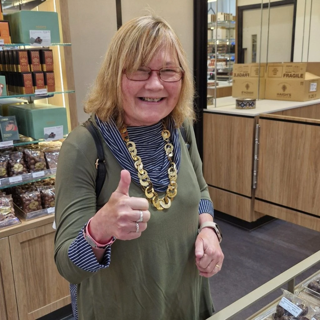 A woman wearing glasses and a large necklace gives a thumbs up inside a shop with shelves and boxes in the background.