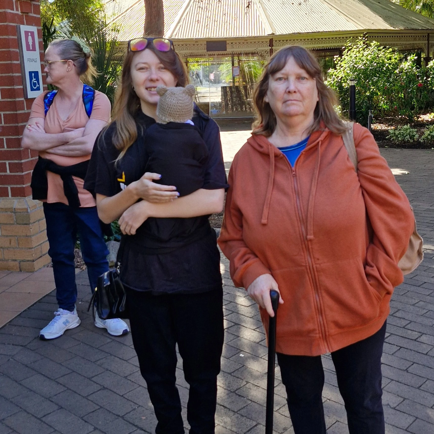 Three women stand outdoors; one holds a small monkey, another uses a cane, and the third stands in the background near a building with signs.