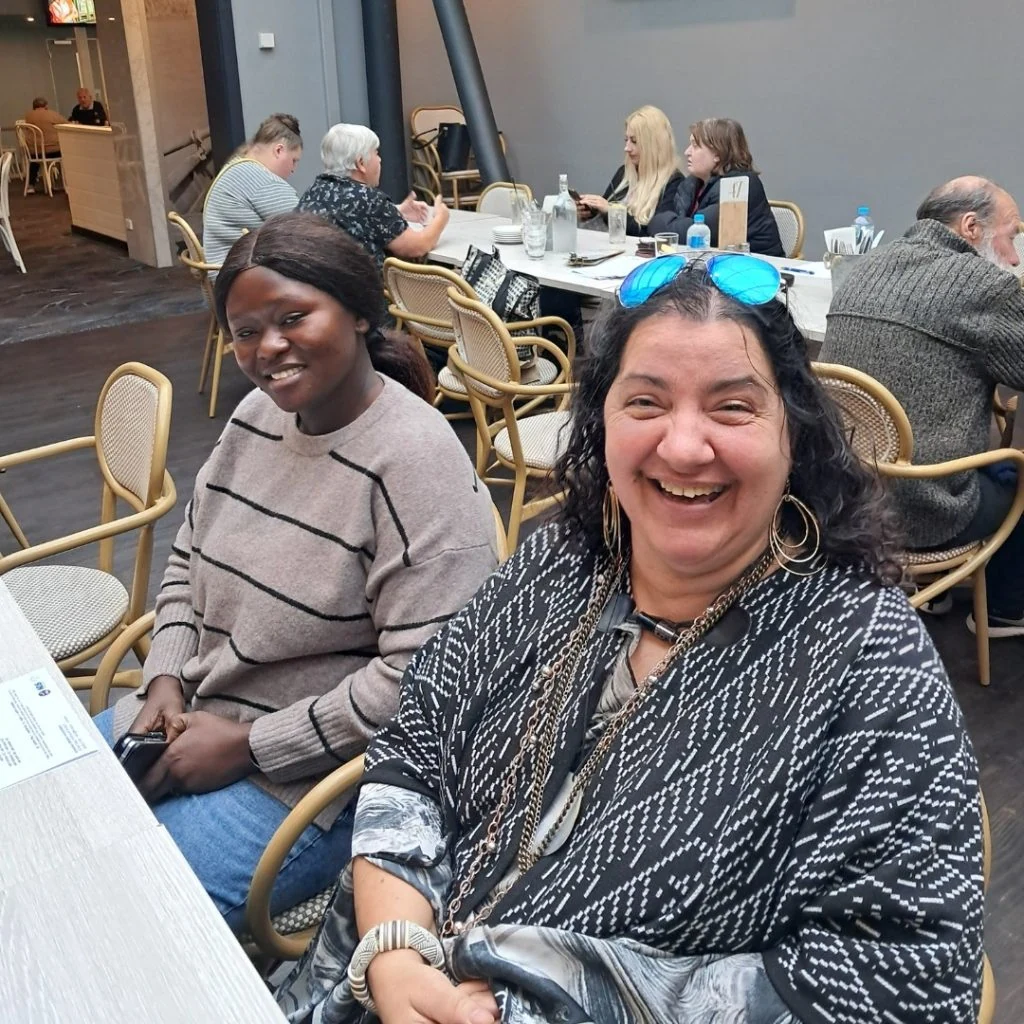 Two women are seated at a table inside a restaurant, smiling at the camera. Several other diners are seated in the background.