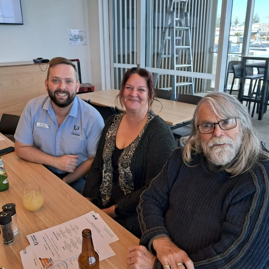 Three people are seated at a table in a restaurant, smiling at the camera. A man in a light blue short-sleeve shirt, a woman in a patterned top, and an older man with long hair wearing glasses.