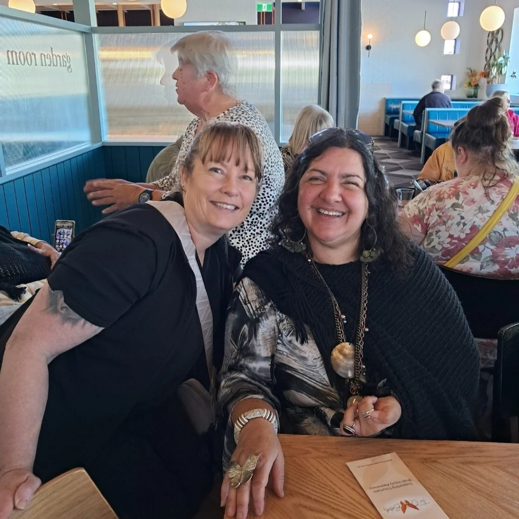 Two women sitting at a dining table, smiling. One woman, in black, leans toward the other woman, who is holding a menu card. Other diners are visible in the background.