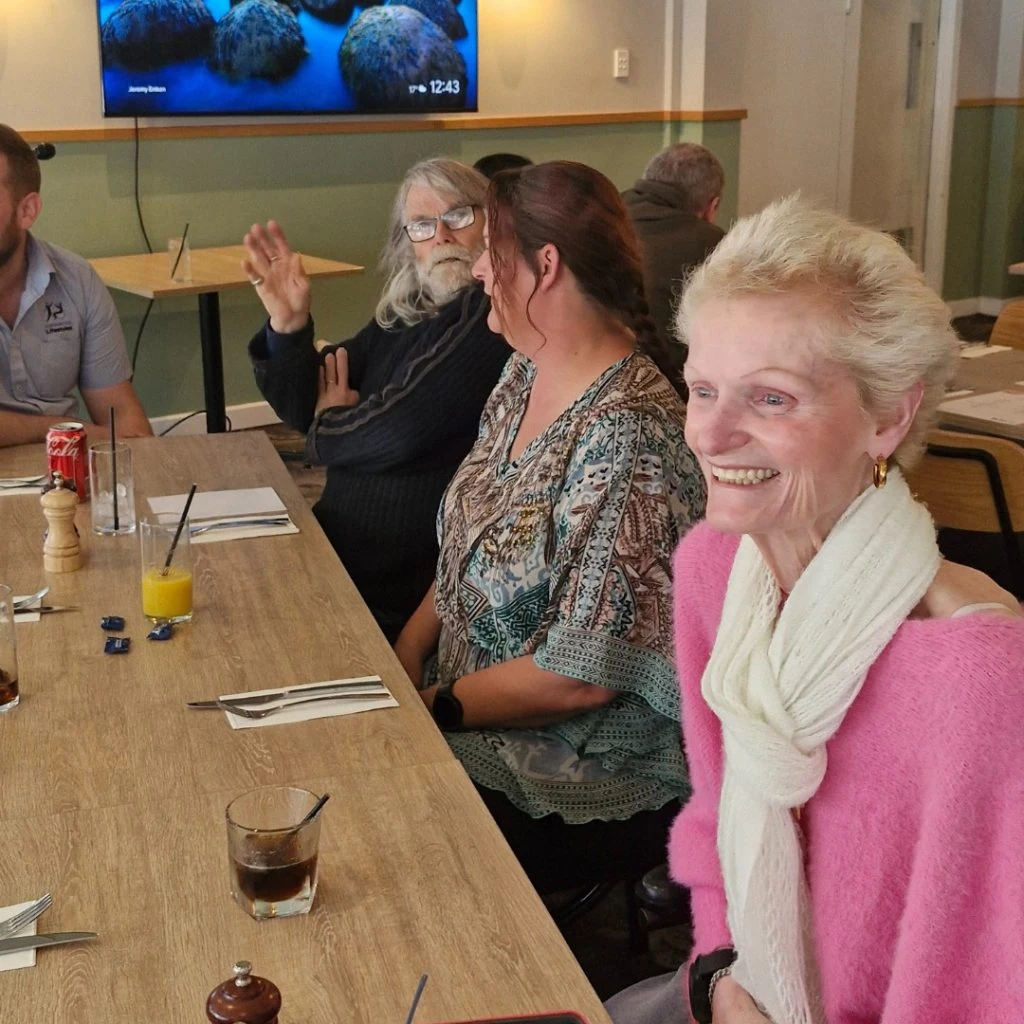 A group of people sitting at a dining table in a restaurant, engaged in conversation and smiling. A TV is on the wall in the background. Drinks and cutlery are on the table.