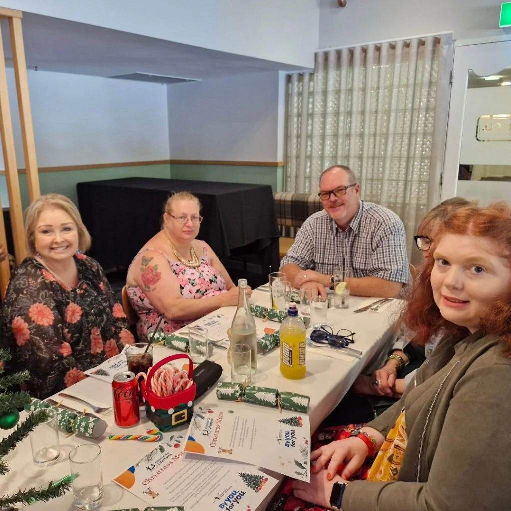 Five people sitting at a table with food and drinks, looking at the camera. Holiday-themed decorations are on the table.