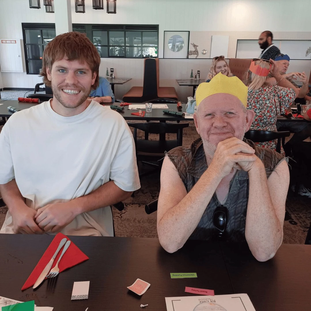 Two people seated at a table, one wearing a yellow paper crown. Other people are in the background at a busy room with tables and decorations.