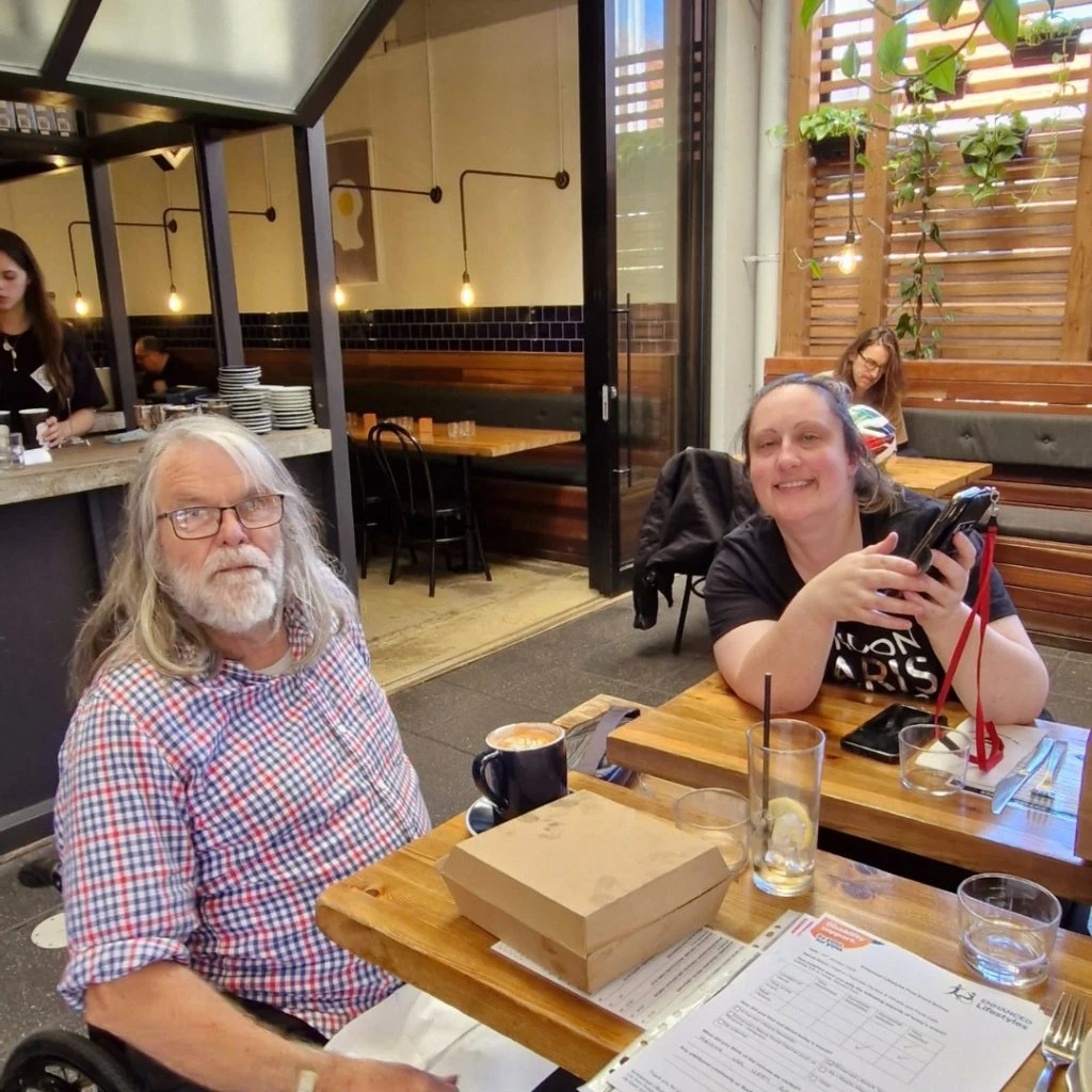 Two people sitting at a café table with drinks and a takeout box; one is holding a phone. There are other patrons and staff in the background.