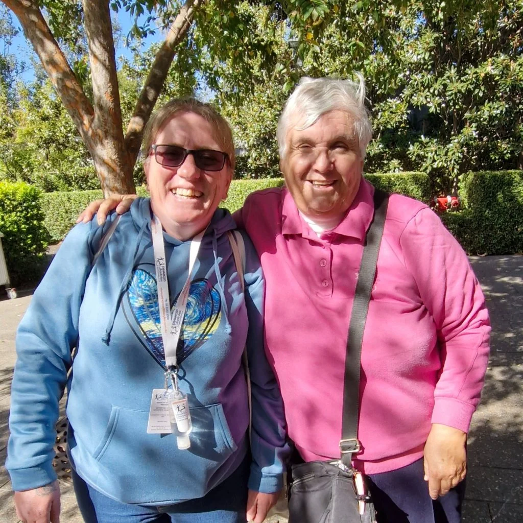 Two women standing outdoors, smiling at the camera. One wears a blue hoodie and sunglasses, the other wears a pink top. Trees and bushes are visible in the background.