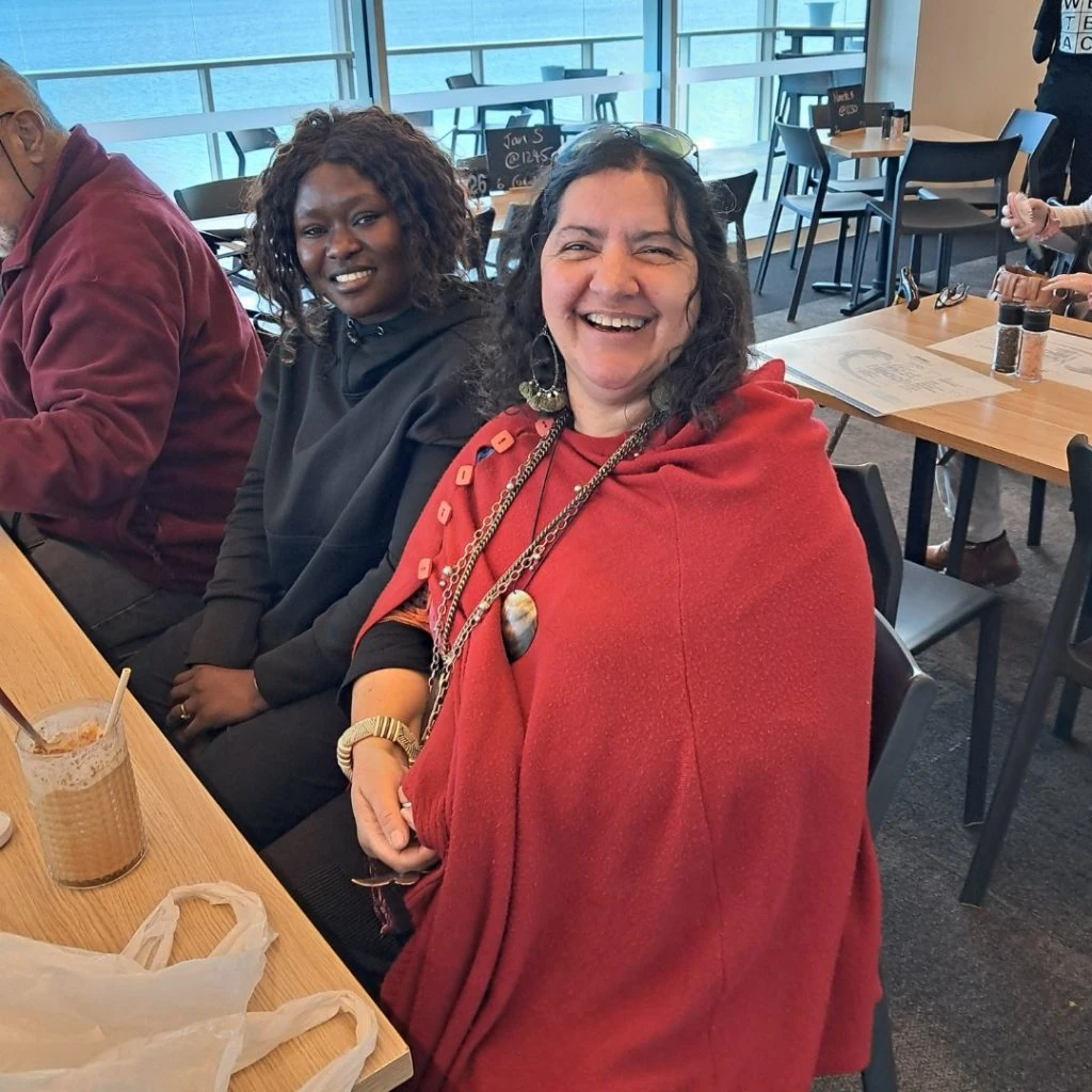 Two women are sitting at a table in a café by the window. The woman on the right is wearing a red shawl and smiling. The woman on the left is dressed in black. Both are seated next to drinks.