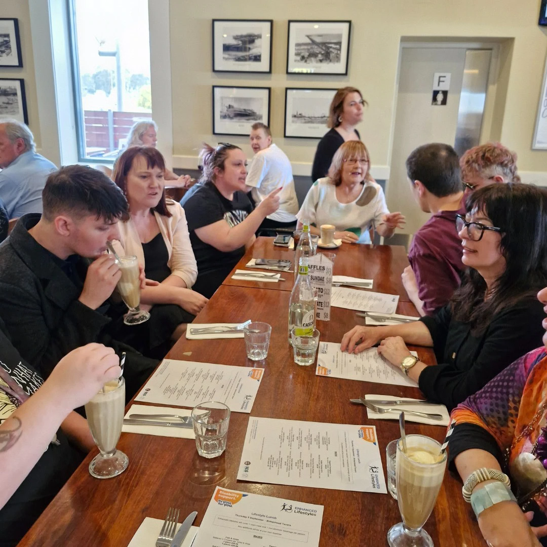 A group of people sitting around a rectangular wooden table in a restaurant, conversing and drinking milkshakes. Menus, cutlery, and a water bottle are on the table.