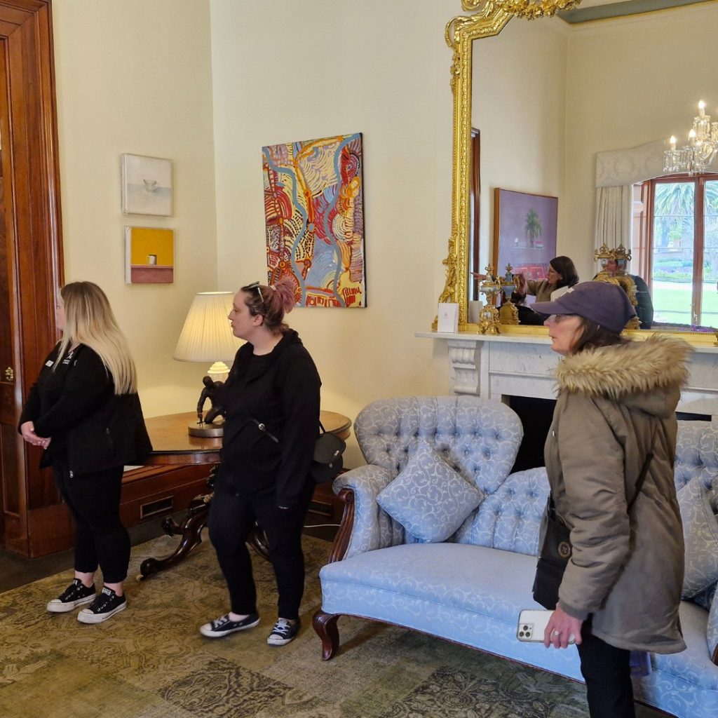 Three people stand inside a room with a blue couch, artworks on the wall, a large ornate mirror, and a chandelier in the background. One person holds a smartphone.