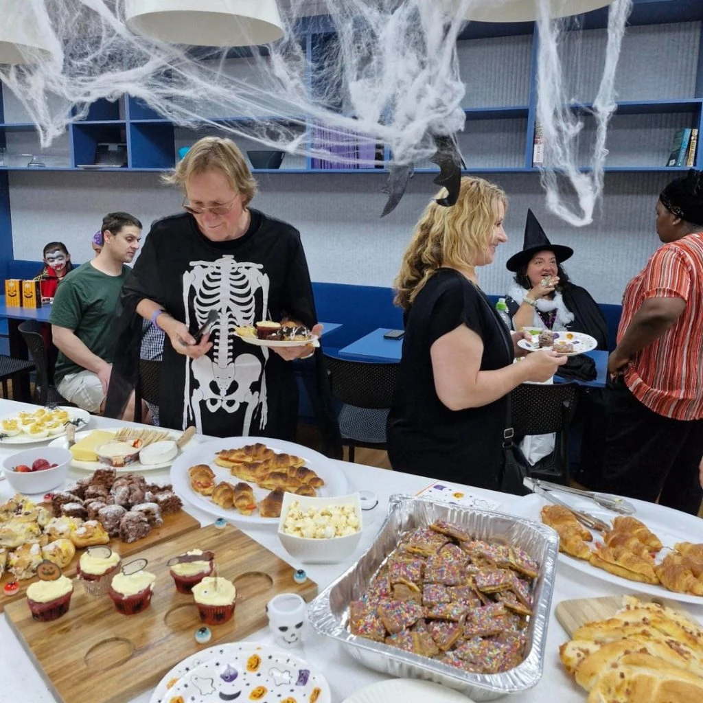 People in Halloween costumes gather around a table filled with various snacks and desserts. The room is decorated with cobwebs.