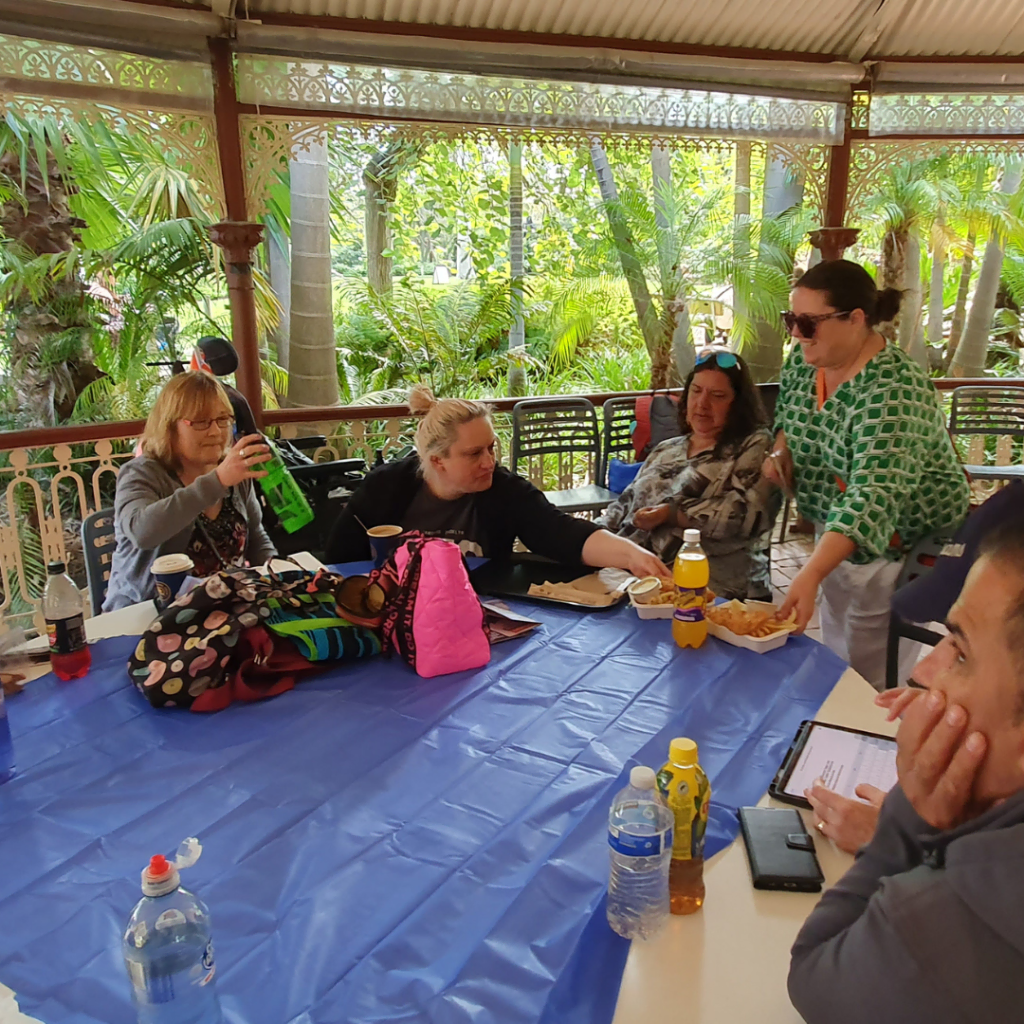 Group of people engaged in conversation at an outdoor table with beverages and personal items.