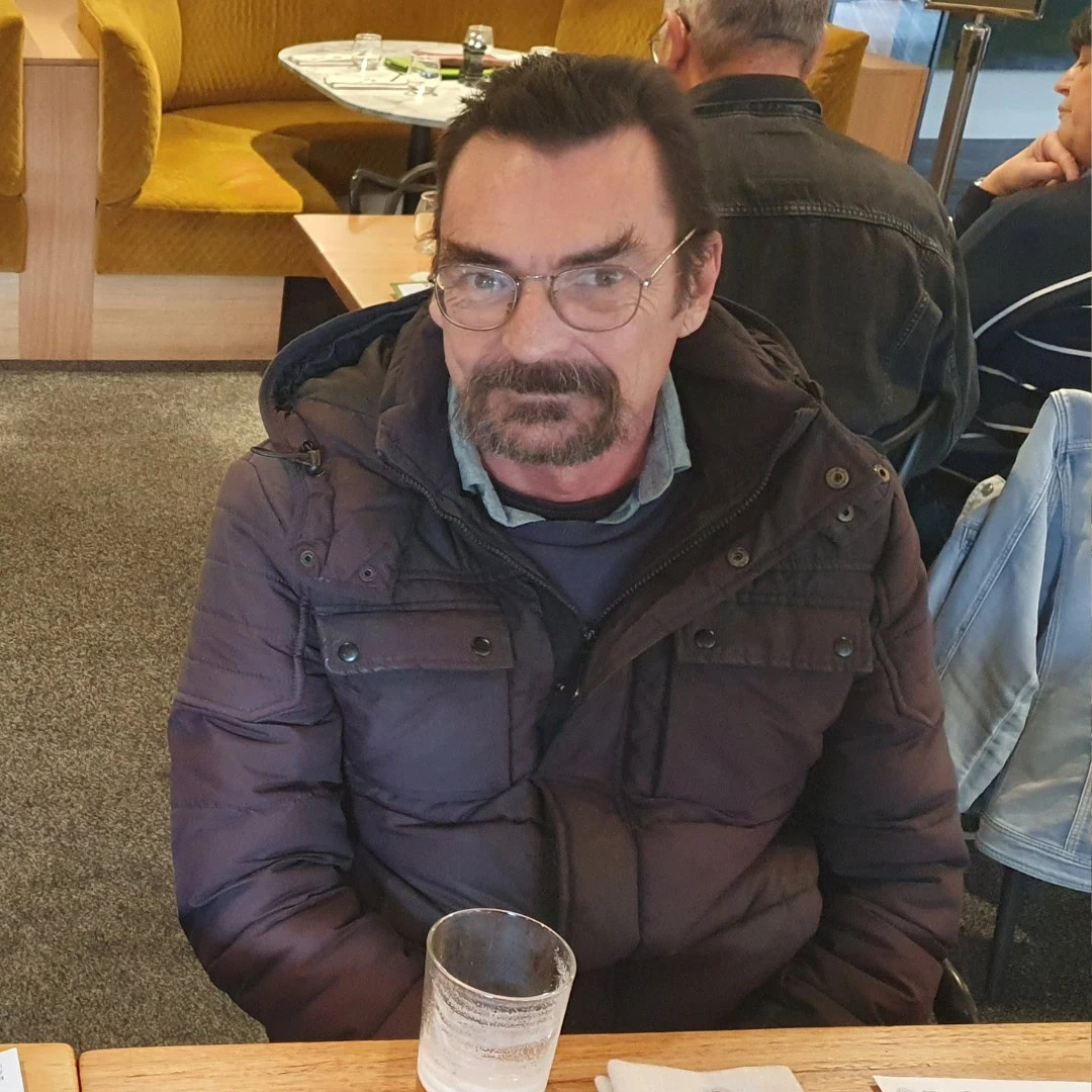 A man with glasses and a dark beard sits at a table in a restaurant, wearing a dark puffy jacket. A nearly empty glass is in front of him.