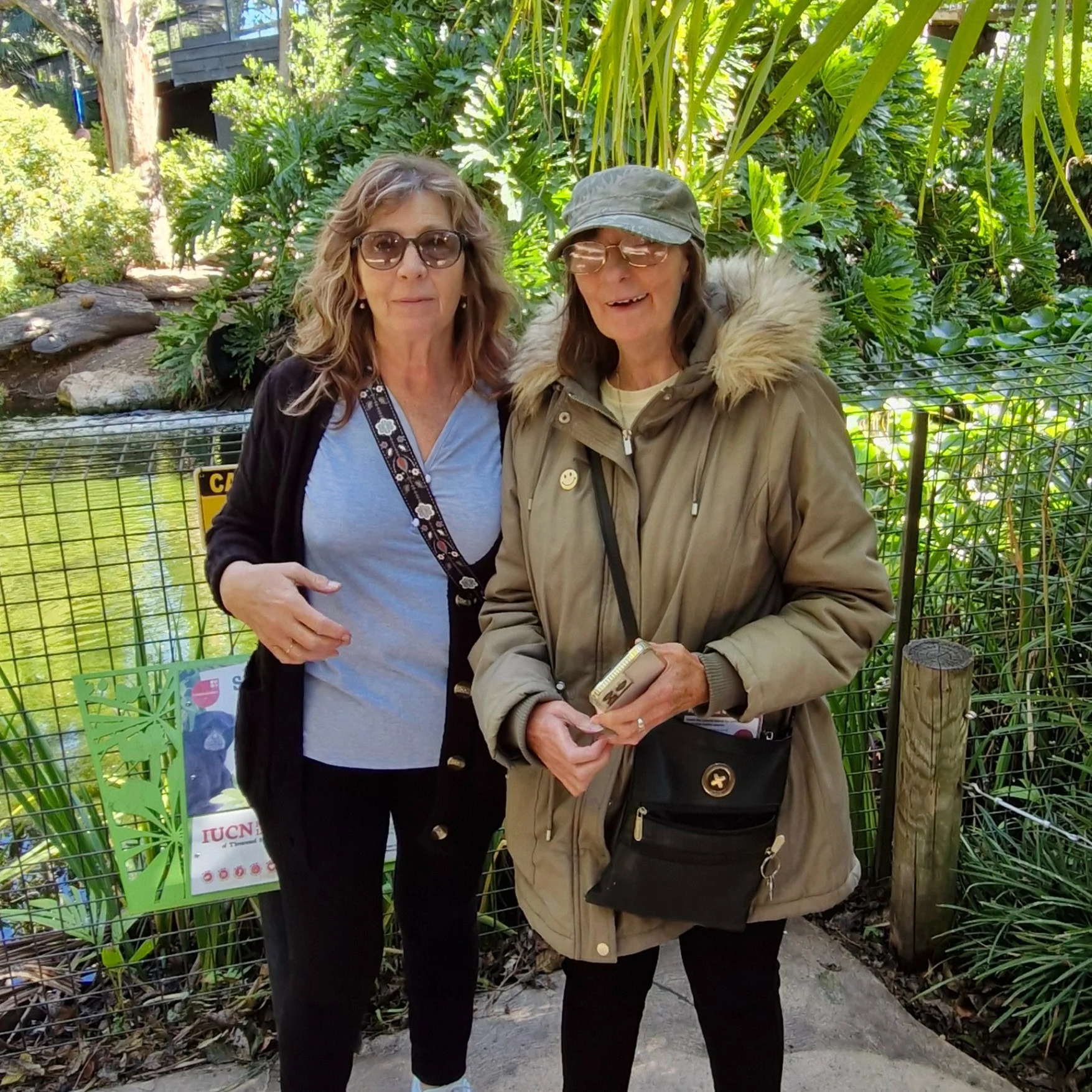 Two women stand in front of a fence and greenery at a park or zoo, both wearing sunglasses and carrying bags. One woman is wearing a coat and cap, the other a gray top.