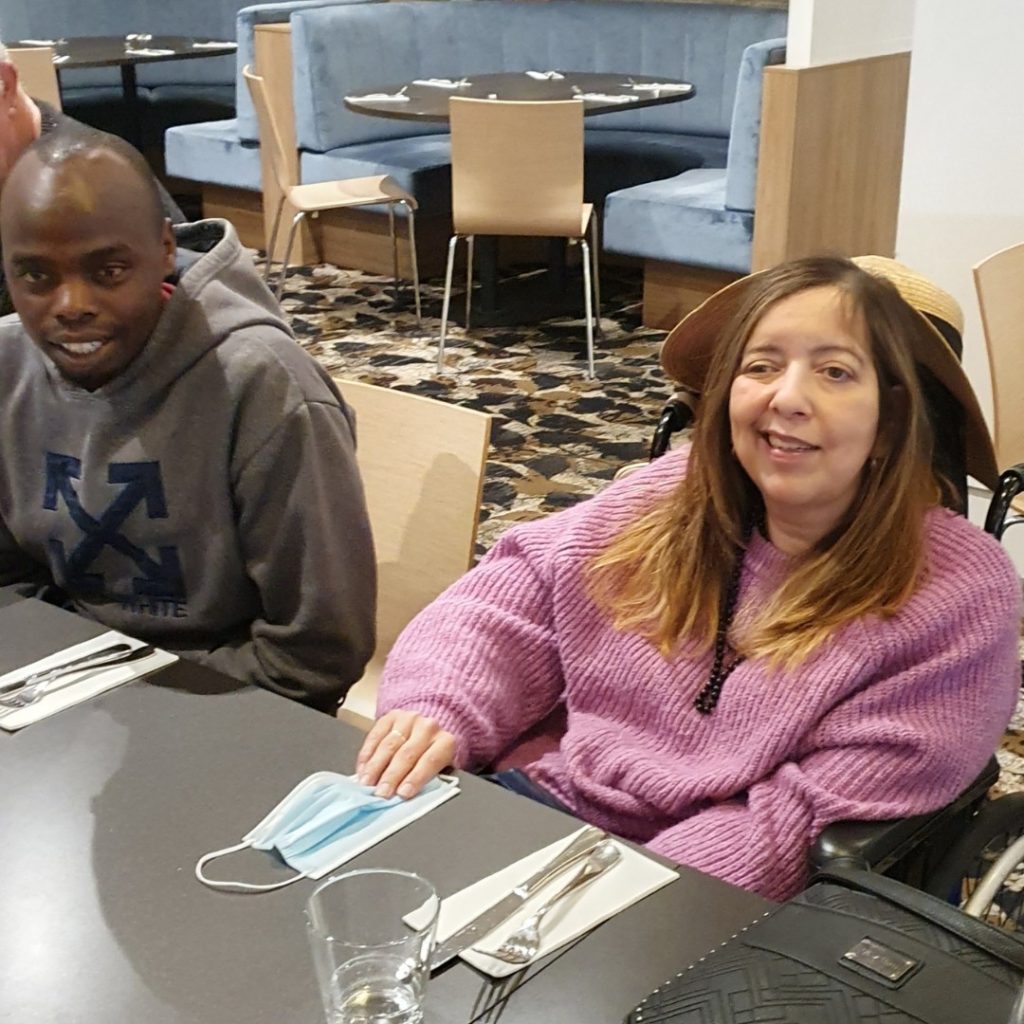 A man and a woman are sitting at a dining table in a restaurant. The woman is in a wheelchair, wearing a purple sweater, and the man is wearing a gray hoodie. A mask and utensils are on the table.
