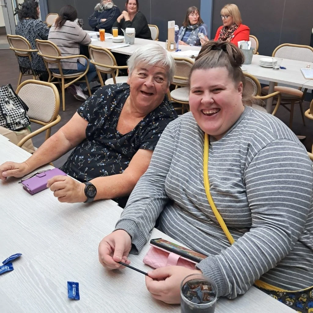 Two people sitting at a table in a casual dining setting, smiling at the camera. Other patrons are seated at tables in the background.