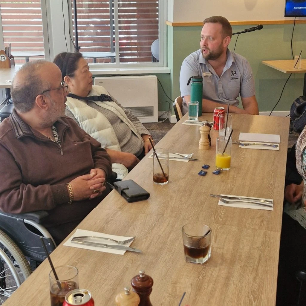 A man in a wheelchair, a woman in a white jacket, and a man in a grey shirt are seated at a table with drinks and cutlery, engaged in conversation at a casual dining setting.