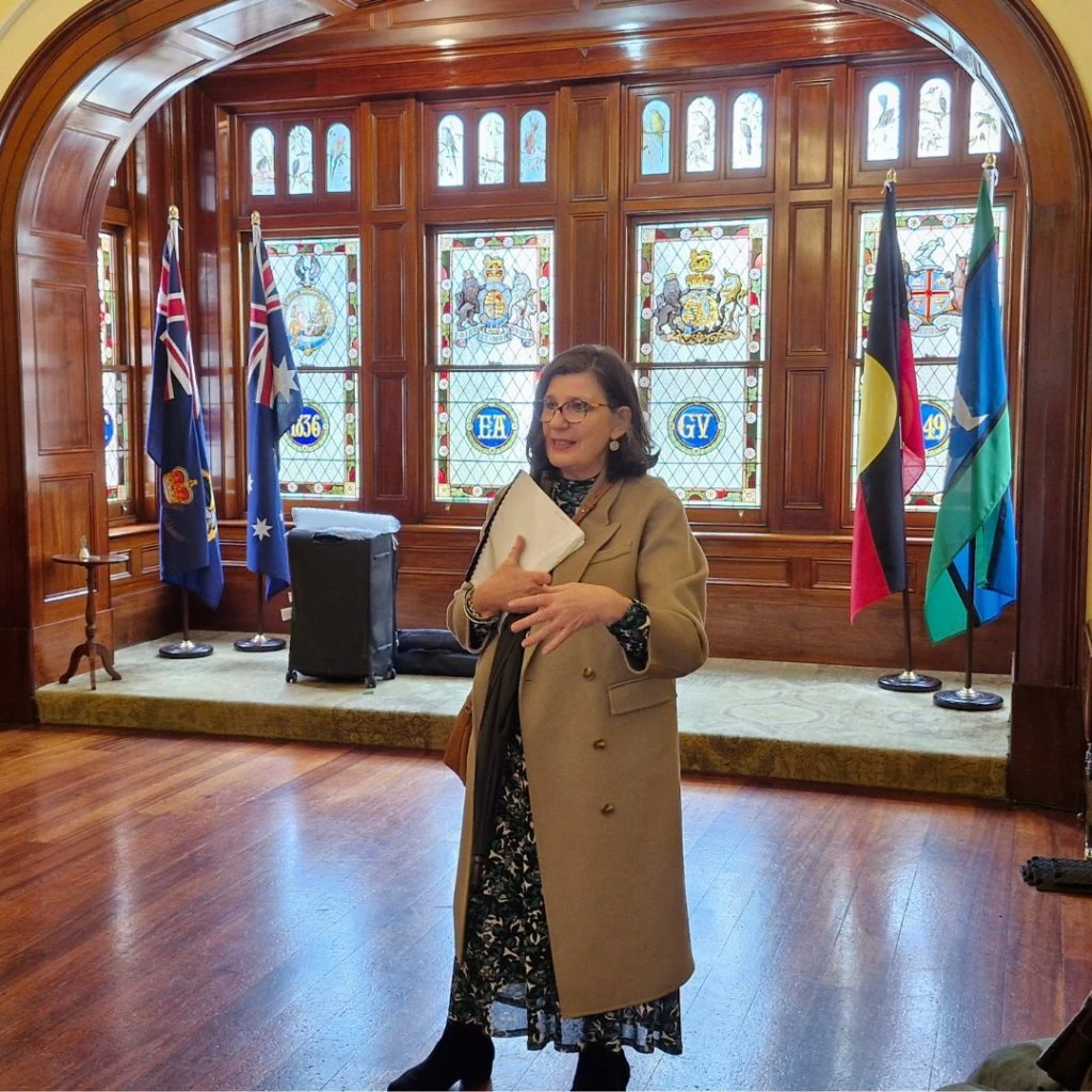 A woman stands in front of stained-glass windows, holding a folder and speaking. Behind her are flags, including the Australian flag, in a room with wood-paneled arches and polished wood flooring.