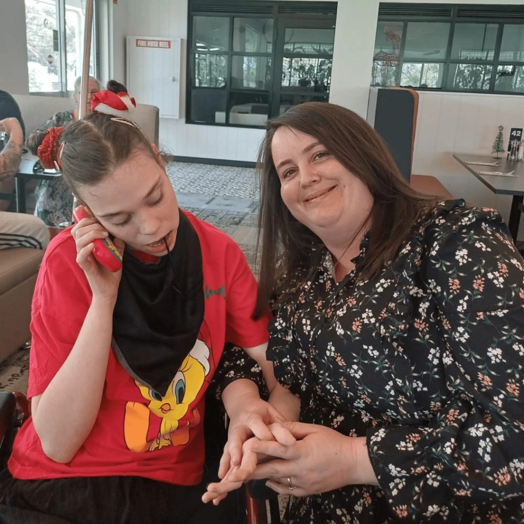 A woman smiles while sitting beside a young person in a red T-shirt who is holding a phone to their ear. They are inside a room with large windows.