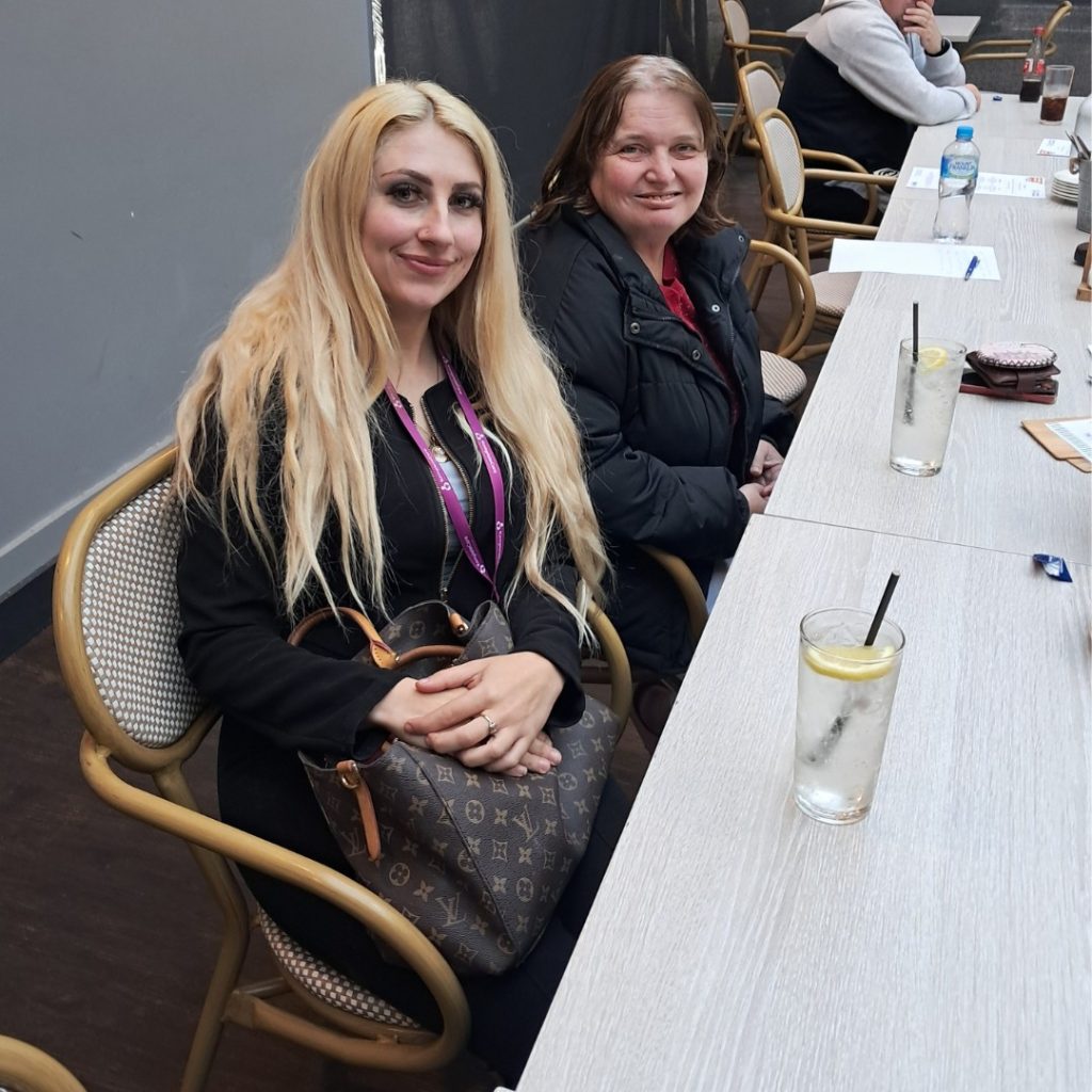 Two women sit at a table with drinks and notebooks in front of them. One has long blonde hair and a Louis Vuitton bag on her lap, while the other has short brunette hair and wears a red-lined black jacket.