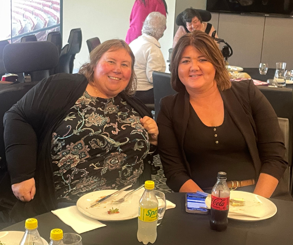 Two women smiling at a table in a stadium.