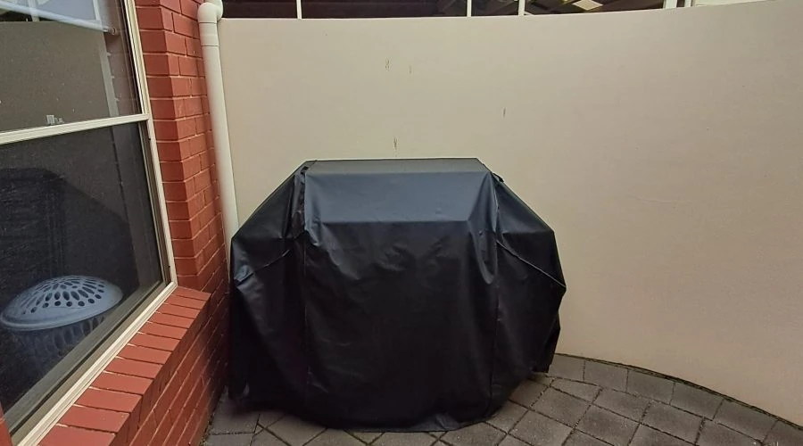 Outdoor corner with a brick wall on the left and a white wall at the back, featuring a covered barbecue grill on a paved patio area.