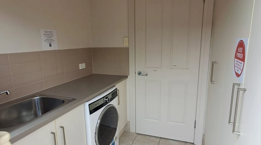 A small laundry room with a sink, a front-loading washing machine, cabinets, and a door. The floor is tiled, and there's a sign on one of the cabinets. The area appears clean and orderly.