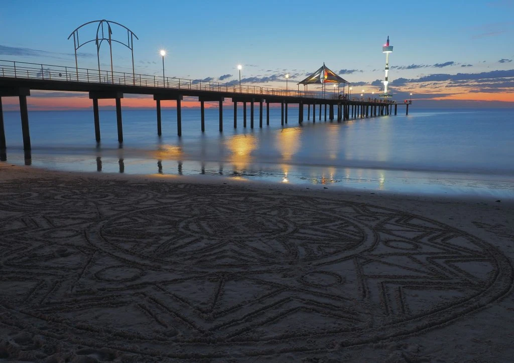 A lit pier extends over calm water at dusk, with an intricate sand design created on the beach in the foreground.