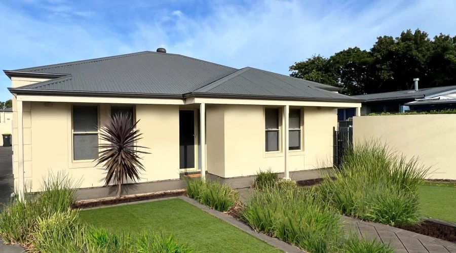 A single-story house with cream-colored walls and a dark gray roof, featuring a small front yard with grass, plants, and a walkway leading to the entrance.