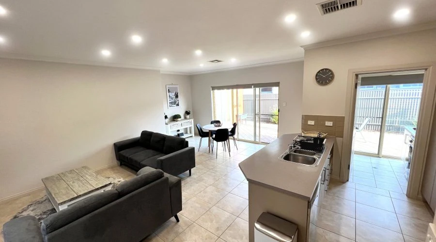 A modern, open-plan living room with grey sofas, a dining table, and a kitchen area. Glass sliding doors lead to an outdoor patio. The room has tiled flooring and recessed lighting.