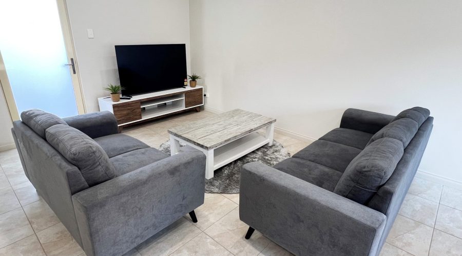 A living room with two gray sofas facing each other, a wooden coffee table in between, and a TV on a media console against the wall. The floor is tiled and there are a few potted plants.