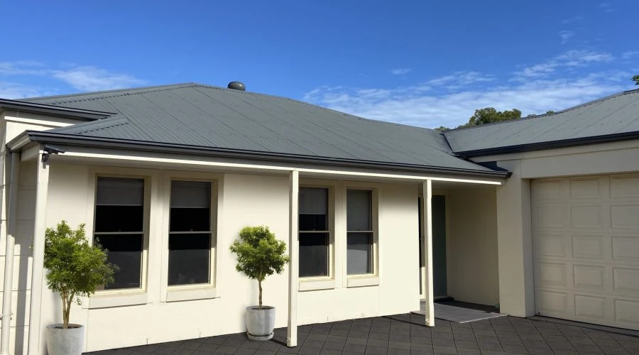 A light-colored, single-story house with a metal roof, two windows, two potted plants, and a garage door, under a clear blue sky.