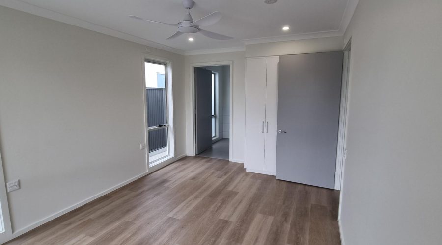 Empty room with light wooden flooring, two windows, a ceiling fan, and a closed gray door leading to another area. Walls are white and plain.