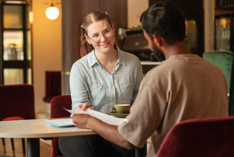 Two people sitting at a table in a cafe, with one person holding a document and the other holding a cup.