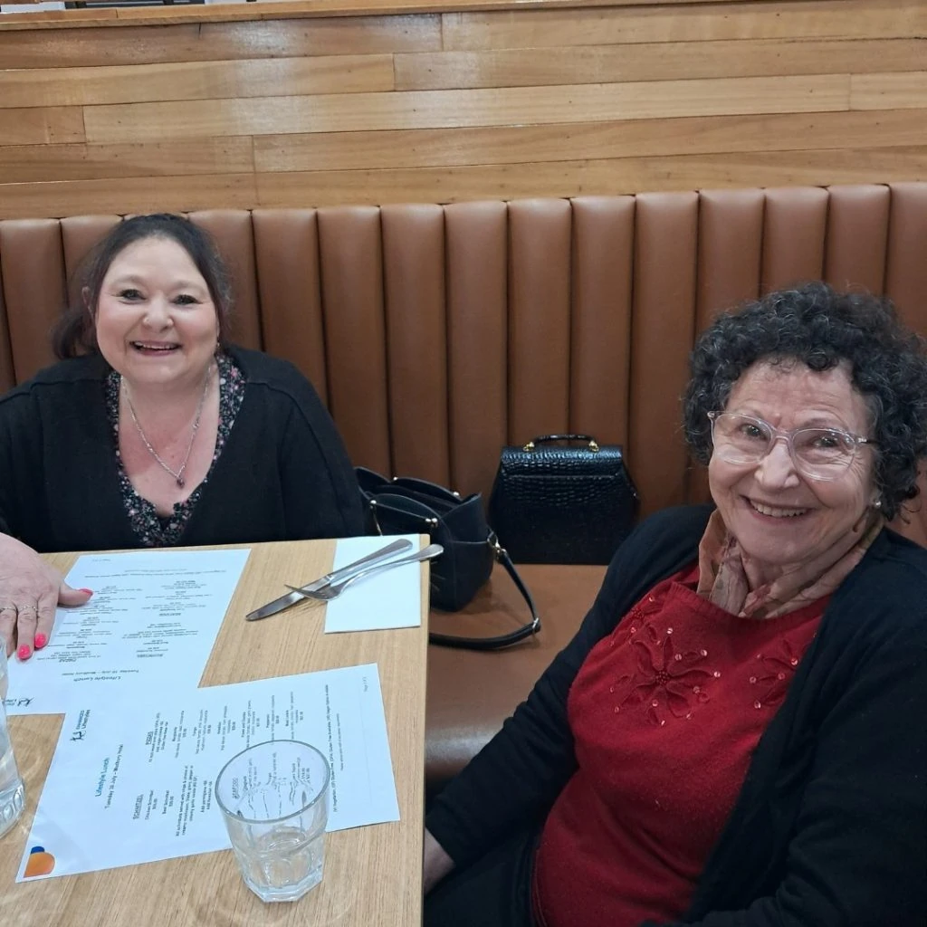 Two women sitting in a restaurant booth, smiling at the camera. Menus, utensils, and glasses are on the table in front of them.