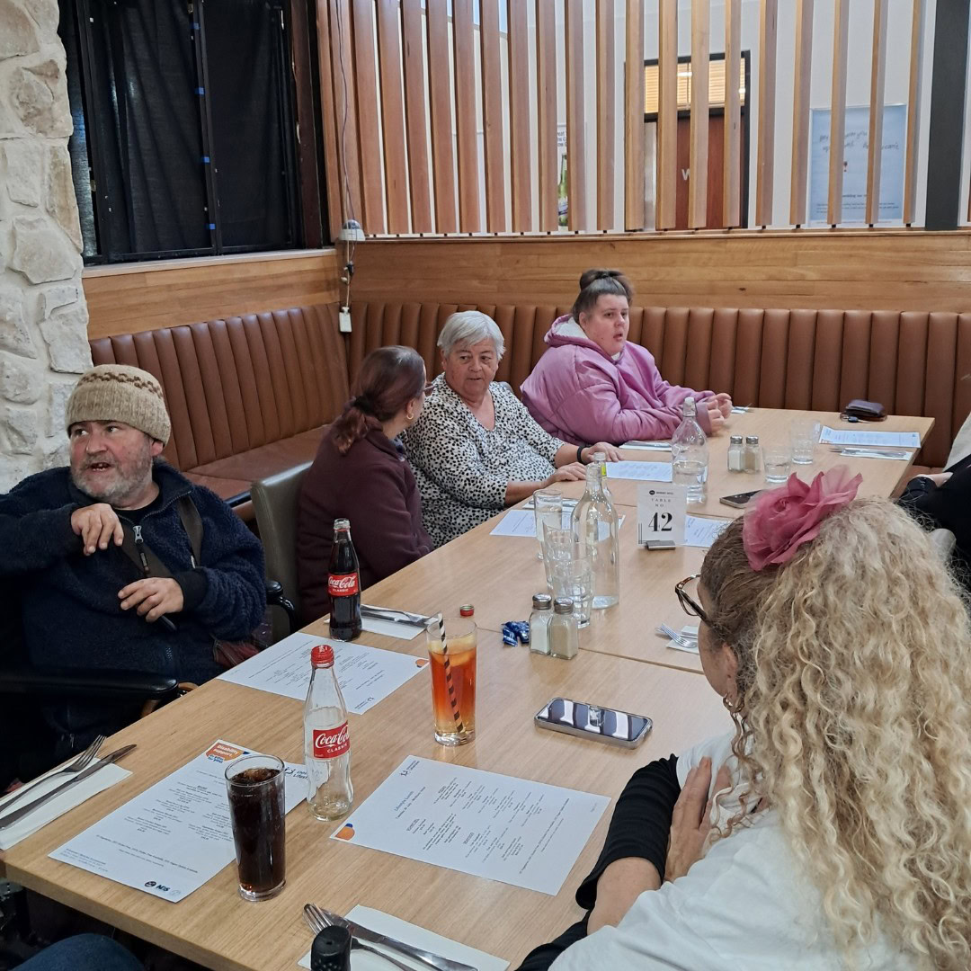 A group of people sit around a rectangular table in a restaurant, engaged in conversation. Various drinks and menus are visible on the table.