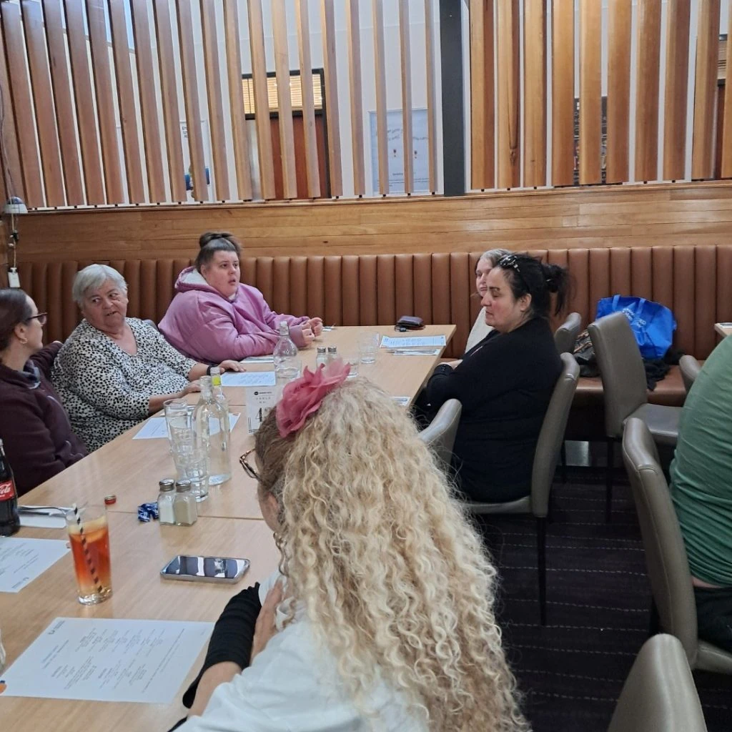 A group of people sit around a wooden table with drinks and papers in a casual meeting room with wood paneling and padded seating. Both men and women are present, some engaged in conversation.