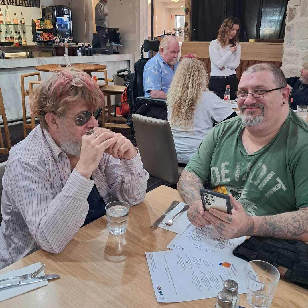 Two men sitting at a restaurant table. One man is wearing sunglasses and the other is holding a smartphone. Two other people and a service counter in the background.