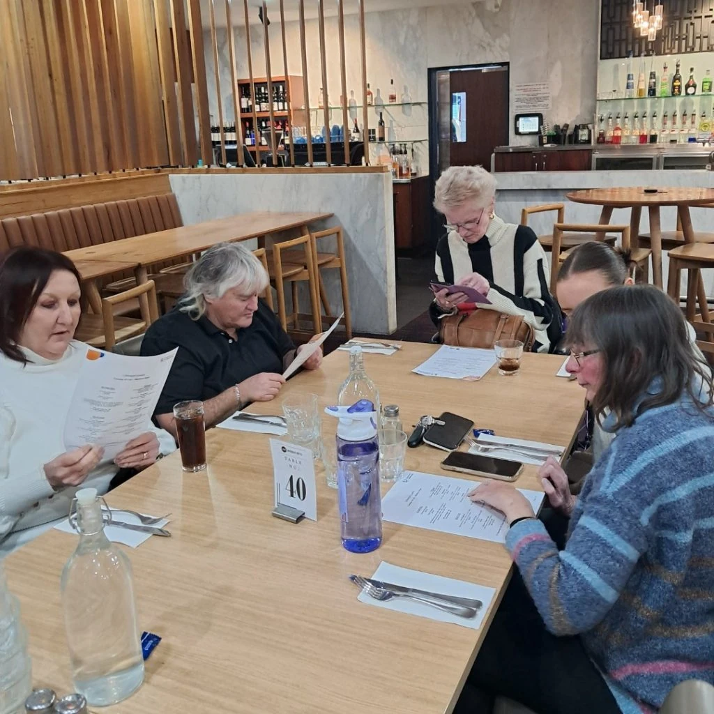 A group of five people sit at a restaurant table studying menus. The table is set with utensils, water bottles, and a number card displaying "40." The background shows a bar area.