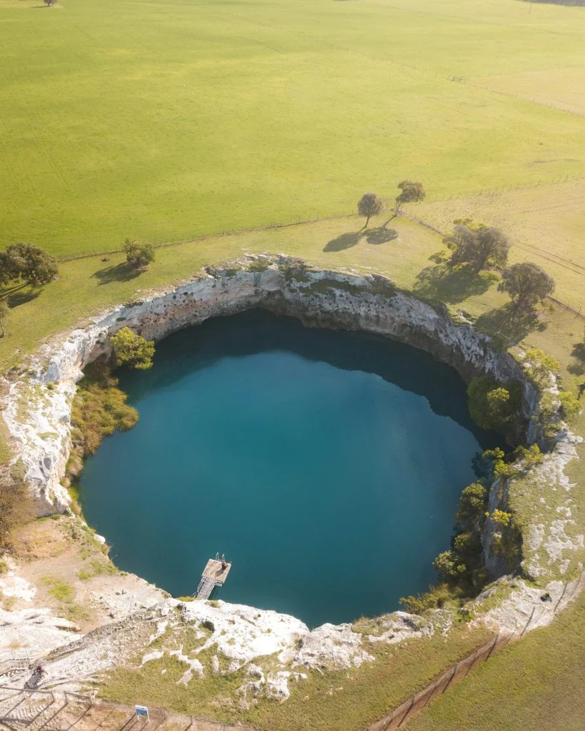 Aerial view of a circular blue water sinkhole surrounded by rocky edges and green fields.