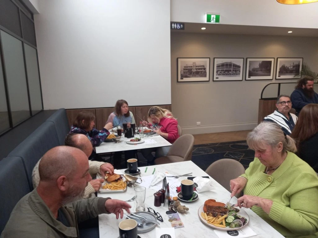 A group of people sitting at tables in a restaurant, eating and engaging in conversation. Plates of food and cups of coffee are on the tables. Framed pictures are on the wall in the background.