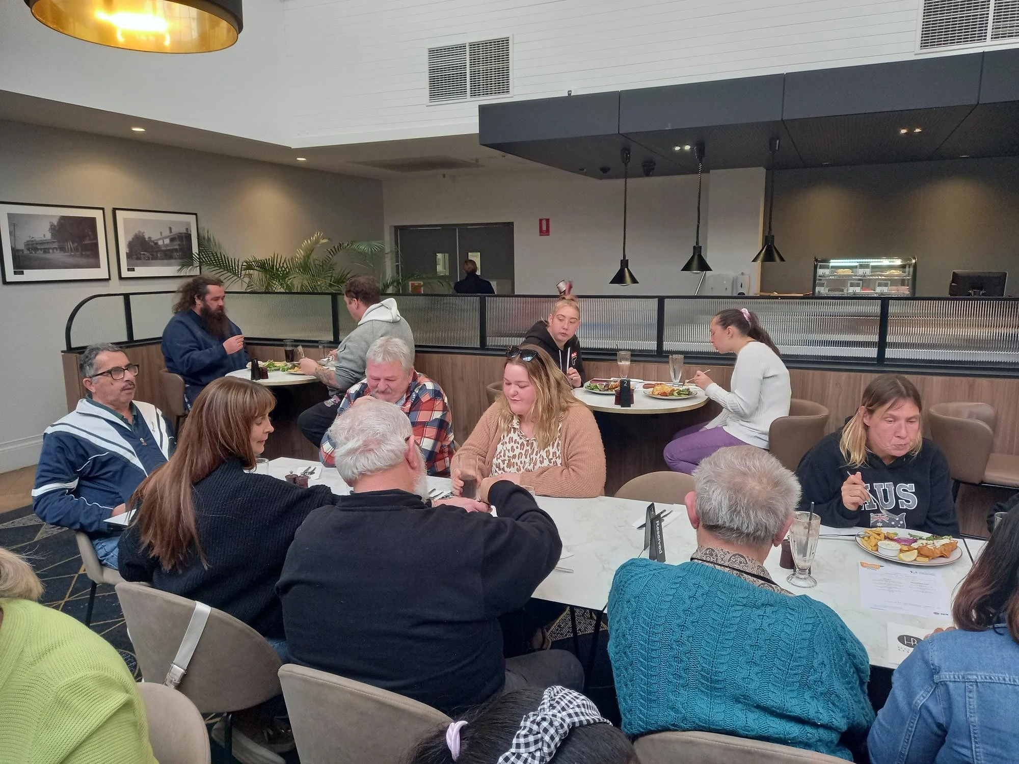 A group of people sitting around tables in a restaurant, eating and talking. Some are looking at menus, while others are engaged in conversation or enjoying their meals.